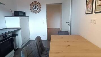 Eine kleine Küche mit einer Mikrowelle, einer Uhr und einem Kühlschrank. Ein Esstisch mit einem Stuhl steht davor.