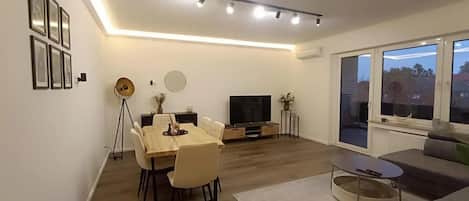A modern living room with a dining area, a flat-screen TV, and a large window.