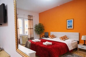 Une chambre d’hôtel avec un lit, une télévision, une fenêtre avec des rideaux, une plante et un tableau au mur.