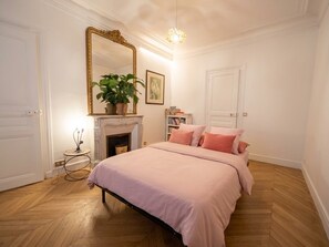 A bedroom with a bed, a fireplace, a mirror, and a small table with a plant. - New Listing (Paris)