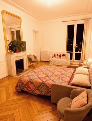 A bedroom with a large bed, a sitting area with a chair, a fireplace, a window with a view of the night sky, and a mirror. - New Listing (Paris)