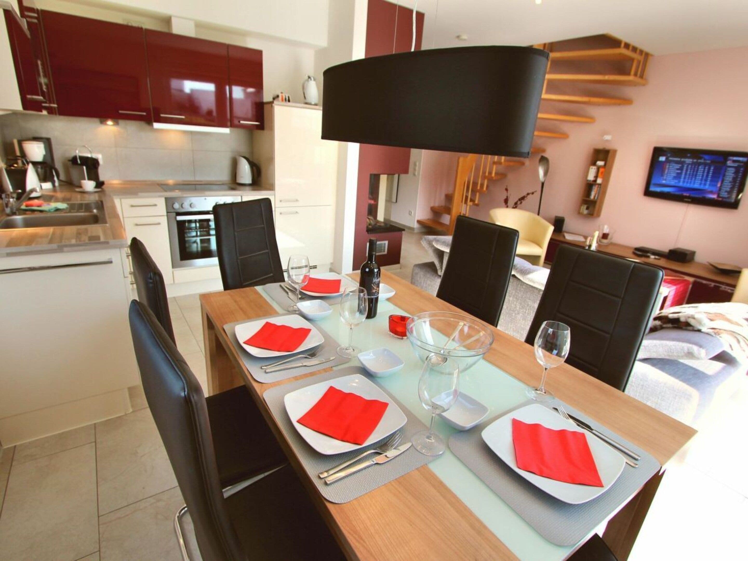 A modern dining area with a long table set for four, equipped with a coffee machine, a kettle, and a television in the background.