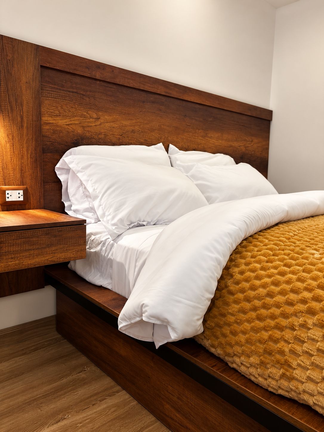 Una cama con ropa de cama blanca y un cabecero marrón, una mesita de noche con toma de corriente y suelo de madera.