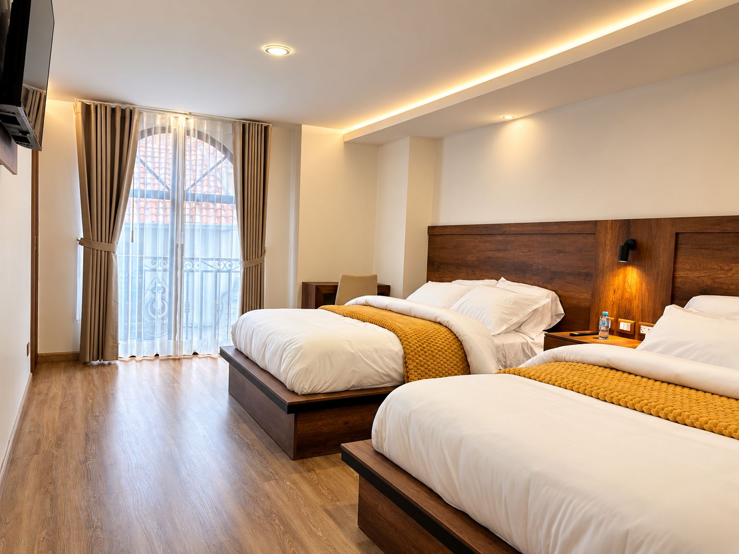 Habitación de hotel con dos camas, cabecero de madera, mesita de noche con lámpara y vista a un edificio a través de una ventana con cortinas.