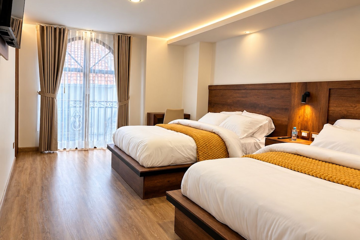 Ein Hotelzimmer mit zwei Betten, einem Holzkopfteil, einem Nachttisch mit Lampe und Blick auf ein Gebäude durch ein fenster mit Vorhängen.
