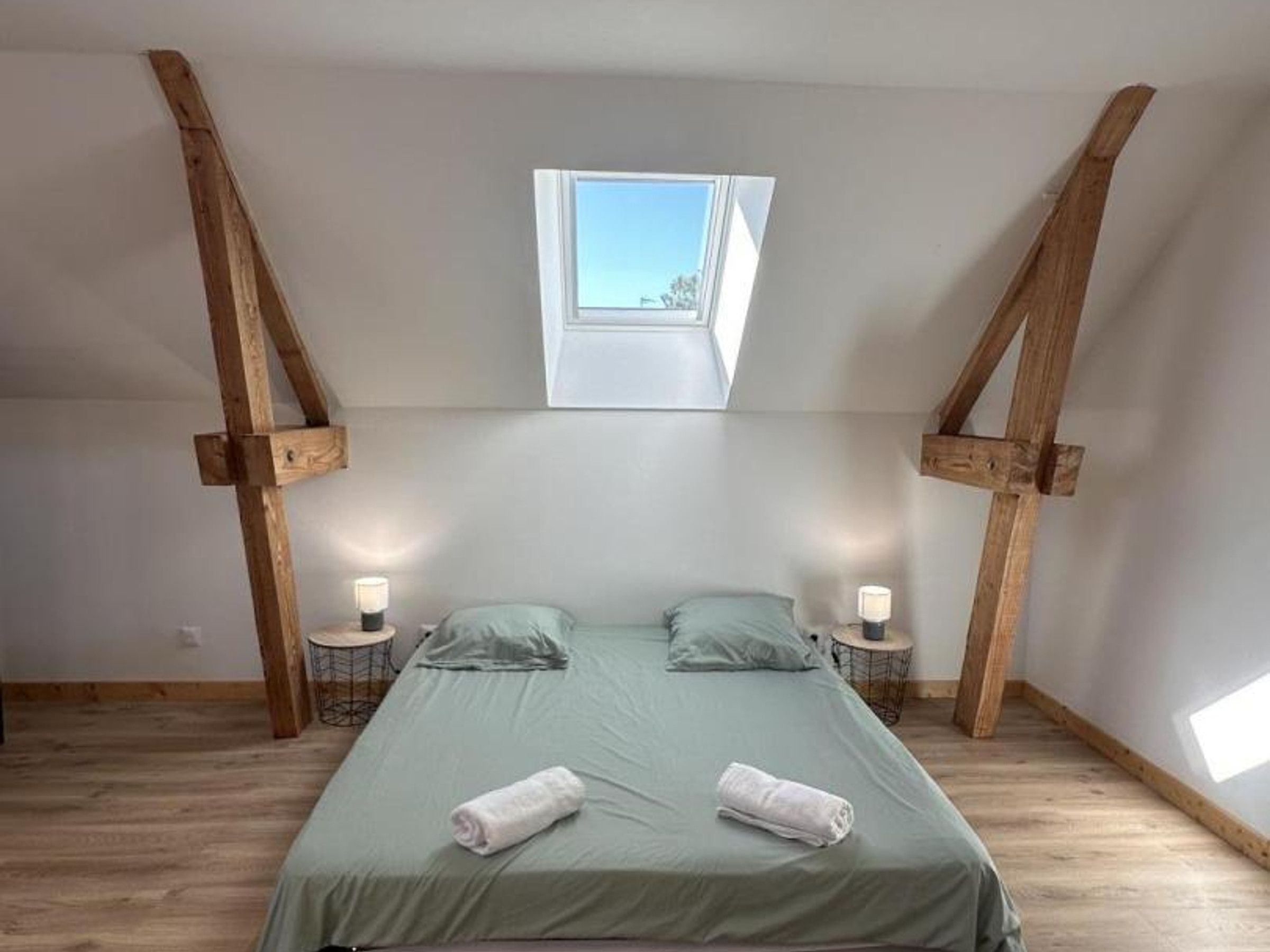 Une chambre avec un plafond en pente, un puits de lumière, deux tables de chevet avec des lampes, un lit avec une literie verte et deux serviettes enroulées.