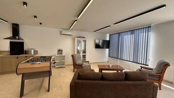 A modern living room with a sofa, a coffee table, a foosball table, a TV, and a window with blinds.