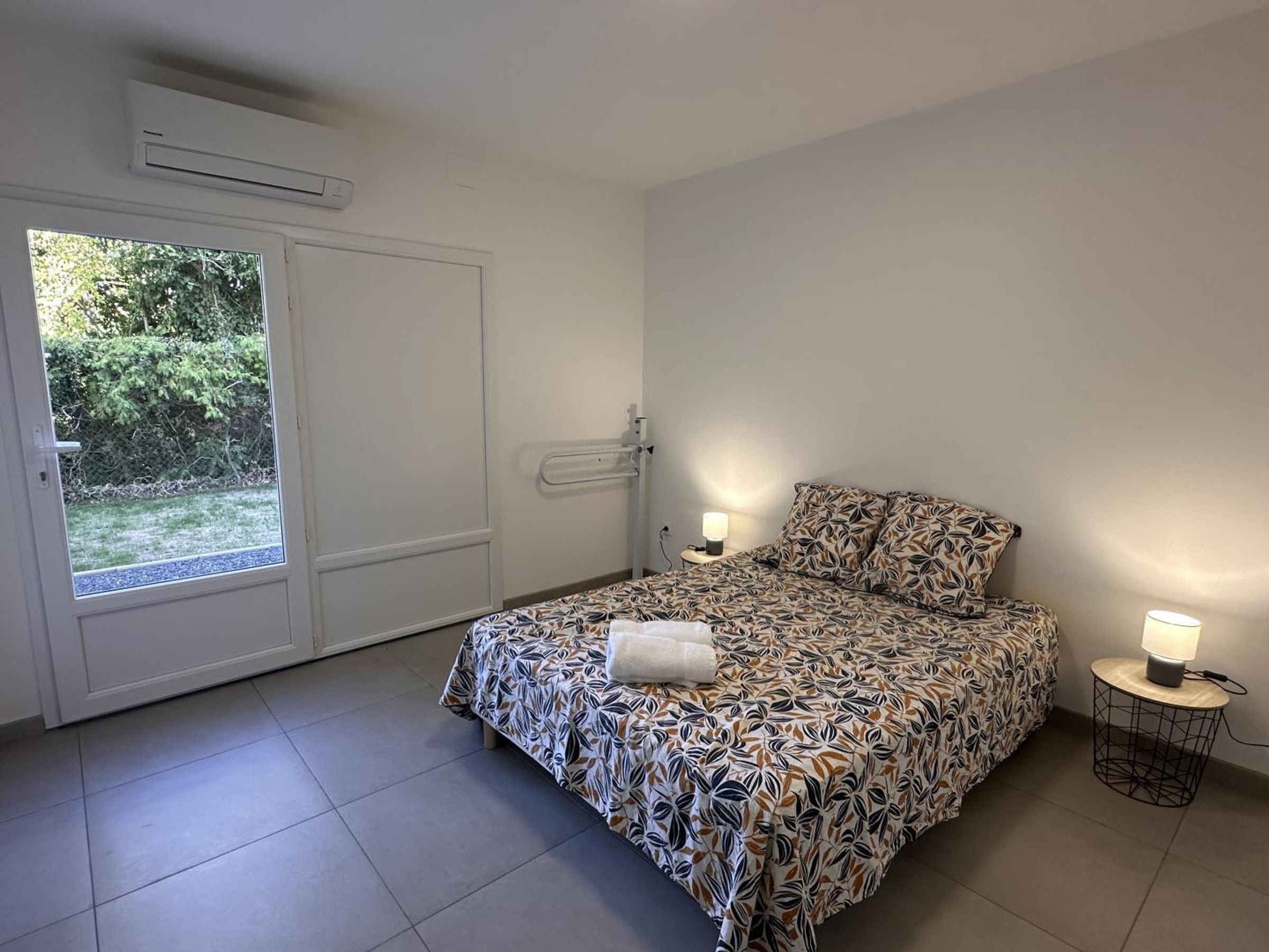 A bedroom with a bed, bedside tables, a door, and a view of greenery outside.