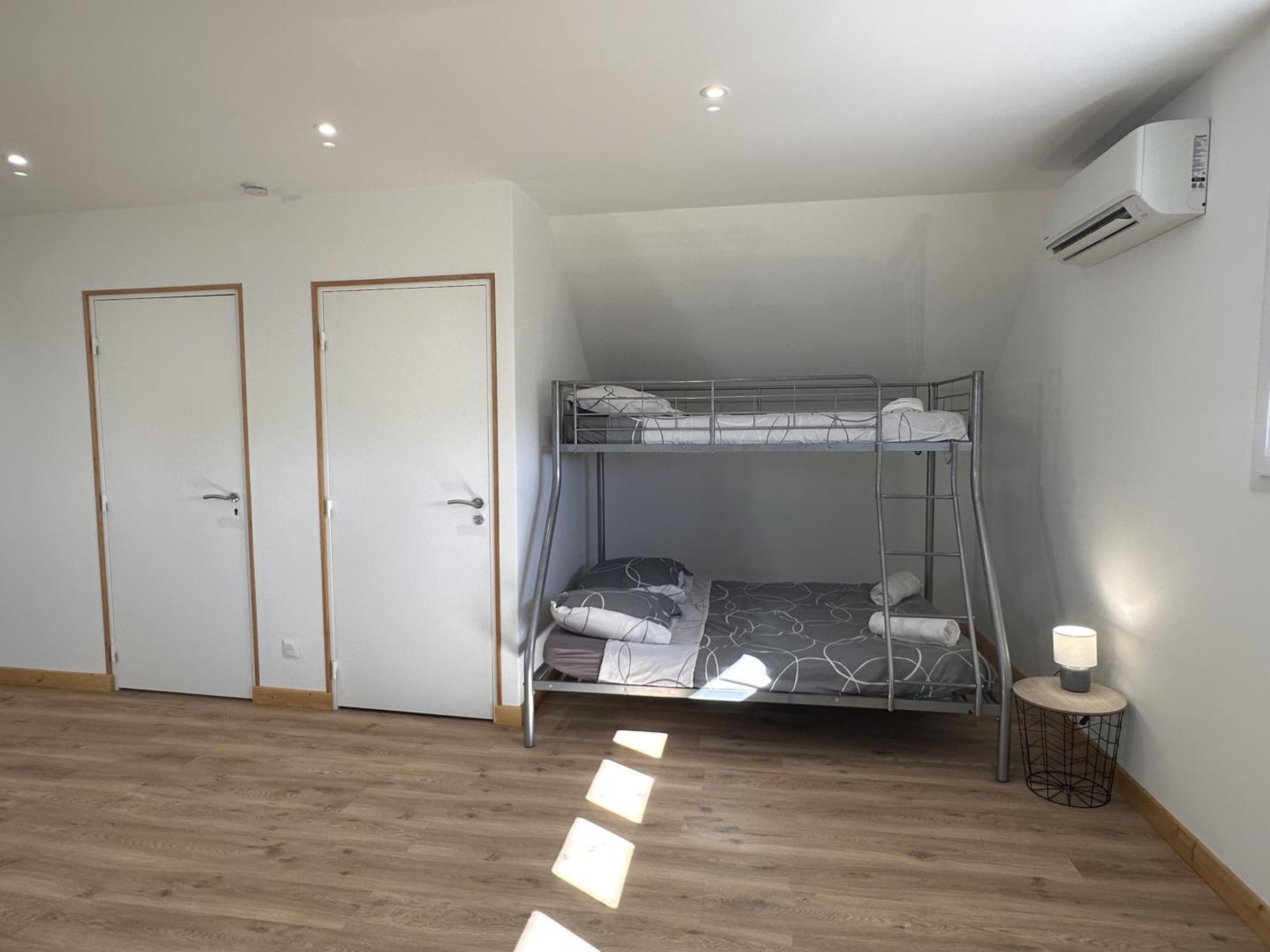 Een kamer met een stapelbed, twee deuren, een klein tafeltje met een lamp en een airconditioning.