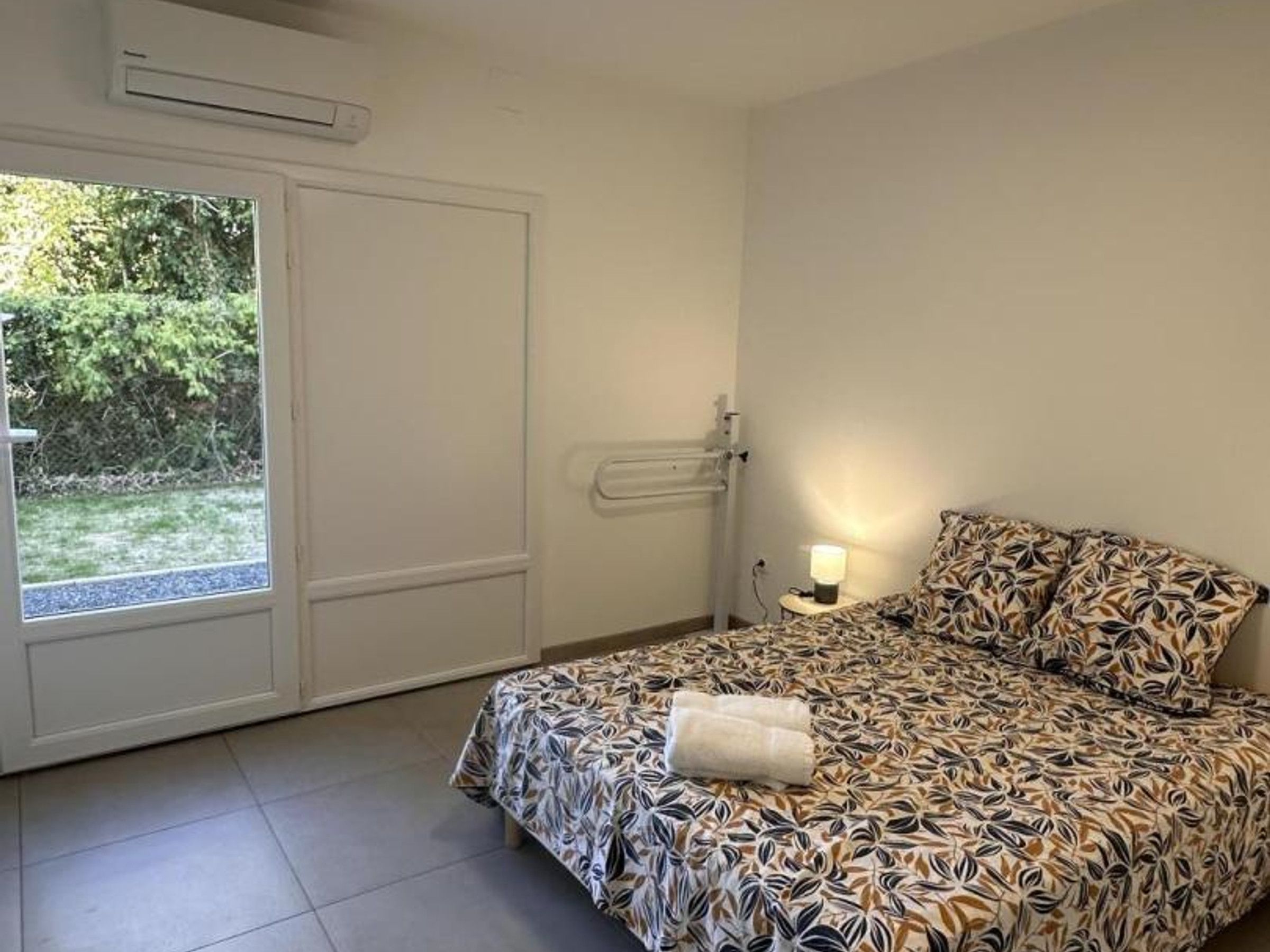 A bedroom with a bed, a bedside table with a lamp, a door, and a view of greenery outside.