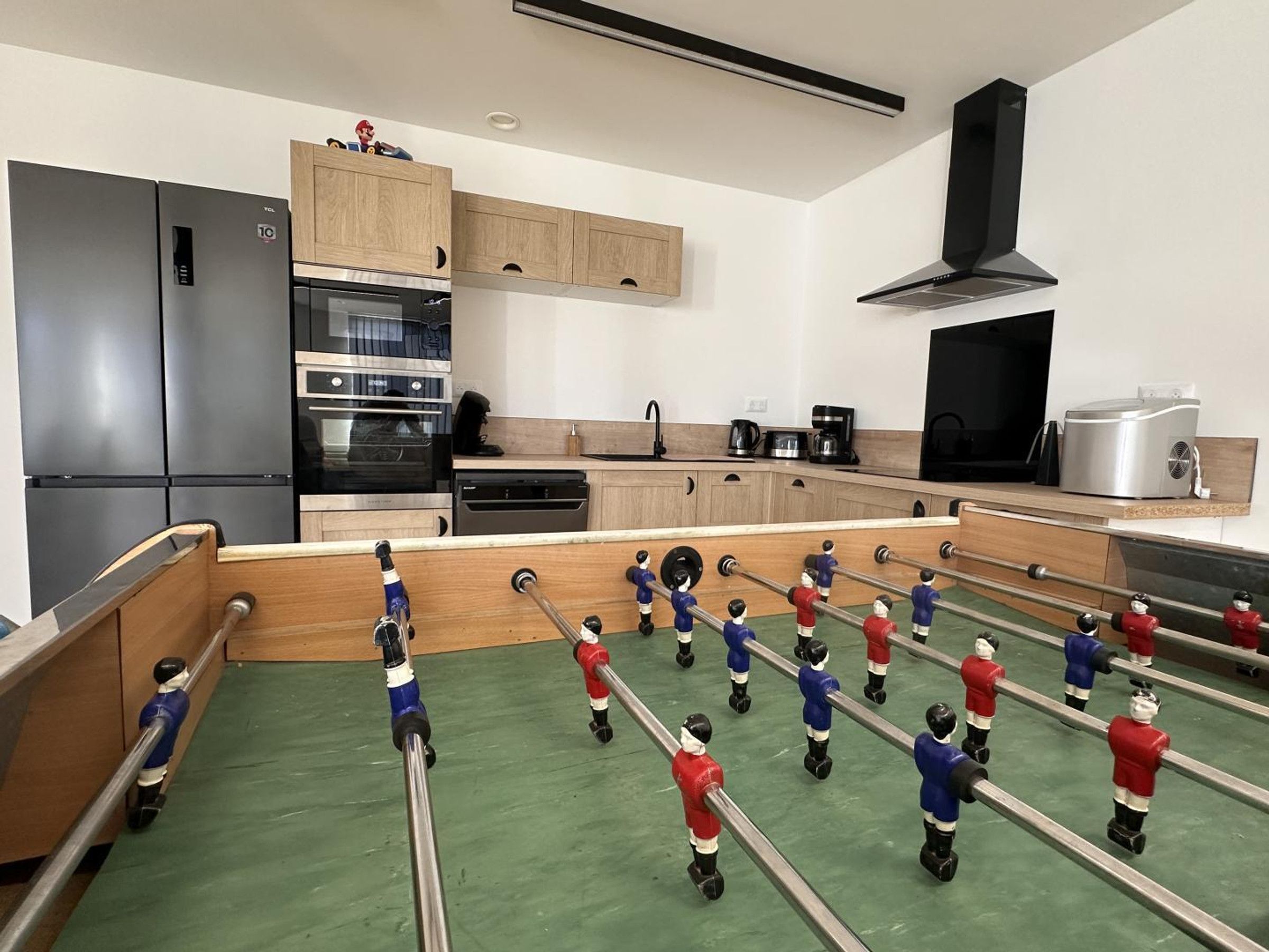 Een keuken met een tafelvoetbaltafel, een koelkast, oven en een koffiezetapparaat.