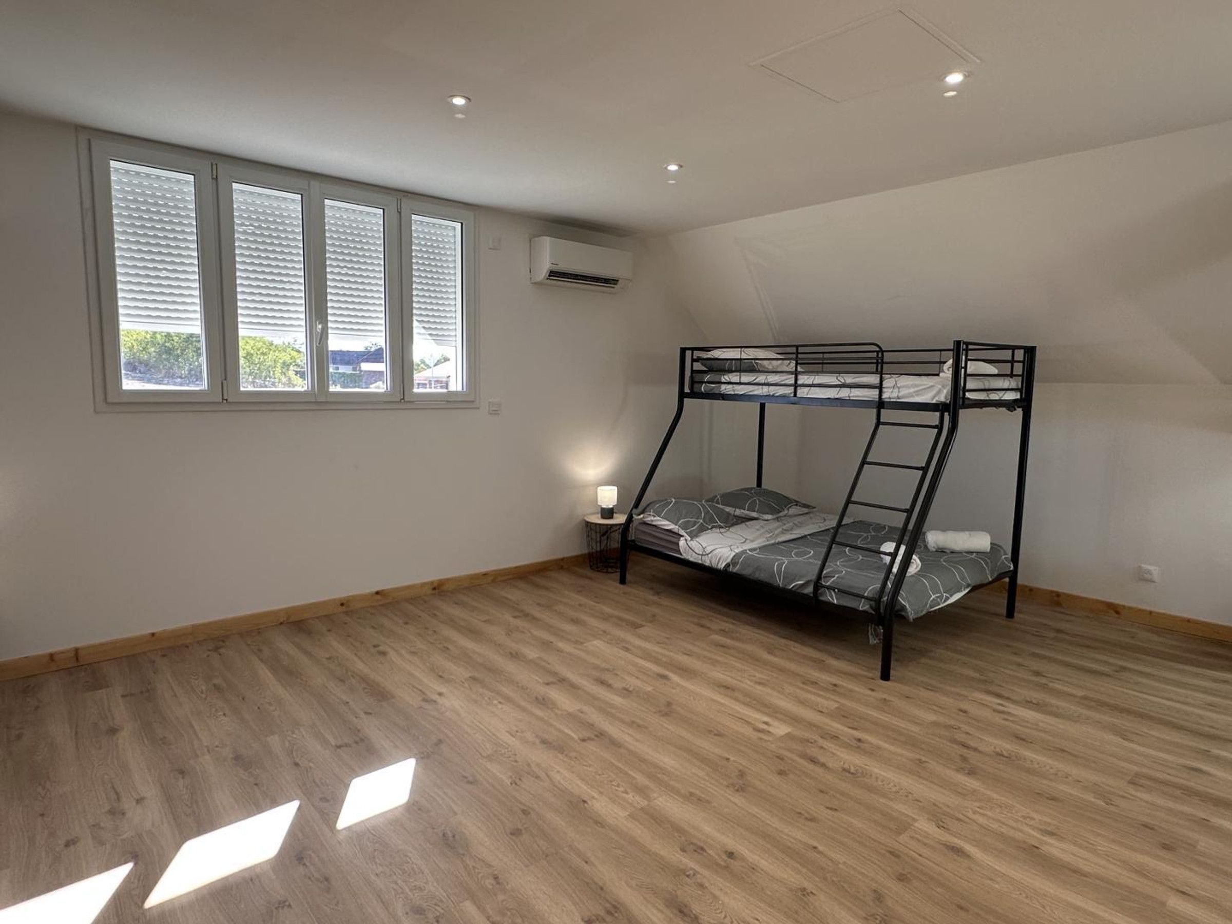 A room with a bunk bed, a window with blinds, an air conditioning unit, and a wooden floor.