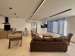 Ein modernes Wohnzimmer mit einer Couch, einem Couchtisch, einem Kicker, einem Fernseher und einem Fenster mit Jalousien.