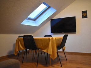 Un comedor con una mesa cubierta con mantel amarillo, sillas negras, un televisor de pantalla plana en la pared y un tragaluz arriba.