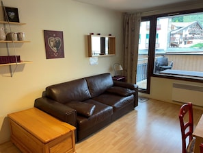 A living room with a brown leather sofa, a wooden side table, a red chair, a small wooden cabinet, a framed picture, and a window with a view of a building. - Studio in the center - 4 guests (Pralognan-la-Vanoise)