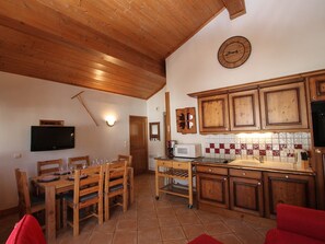 Een houten eettafel met stoelen, een televisie aan de muur, een keuken met kastjes en een magnetron, en een rode bank.