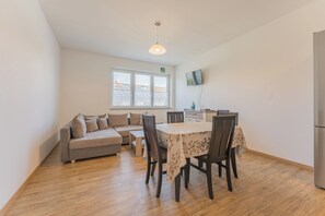 A modern living room with a sofa, a dining table with chairs, a flat-screen TV mounted on the wall, and a window with a view of the roof. - New Listing (Mainz)
