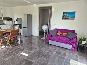 A modern living area with a purple sofa, a dining table with chairs, a kitchen with appliances, and a fan. - New Listing (Sainte-Anne)