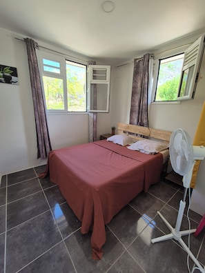 A room with a bed, a standing fan, and windows with curtains. - New Listing (Sainte-Anne)