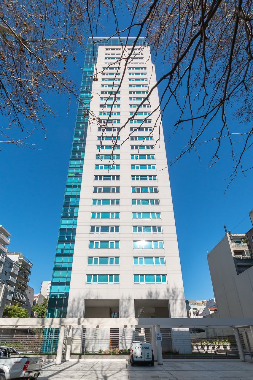 A tall, modern building with multiple floors and large windows, surrounded by other buildings.