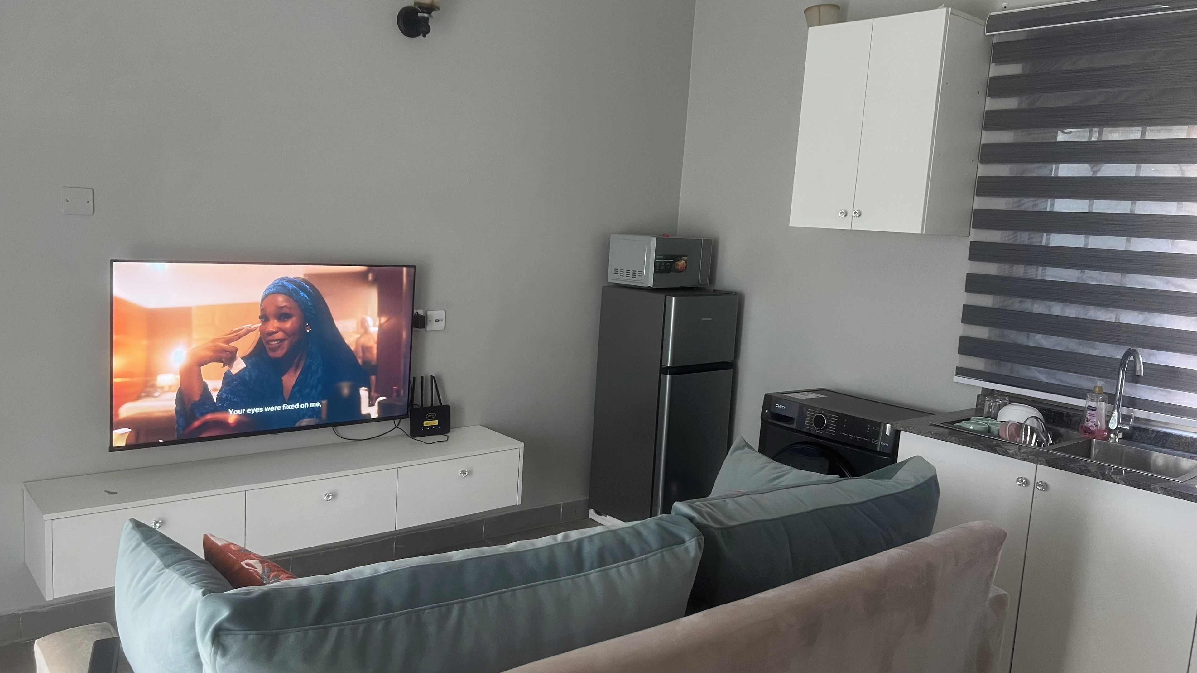 Una sala de estar con televisor, refrigerador, lavadora y un área de cocina con pileta y mostrador.