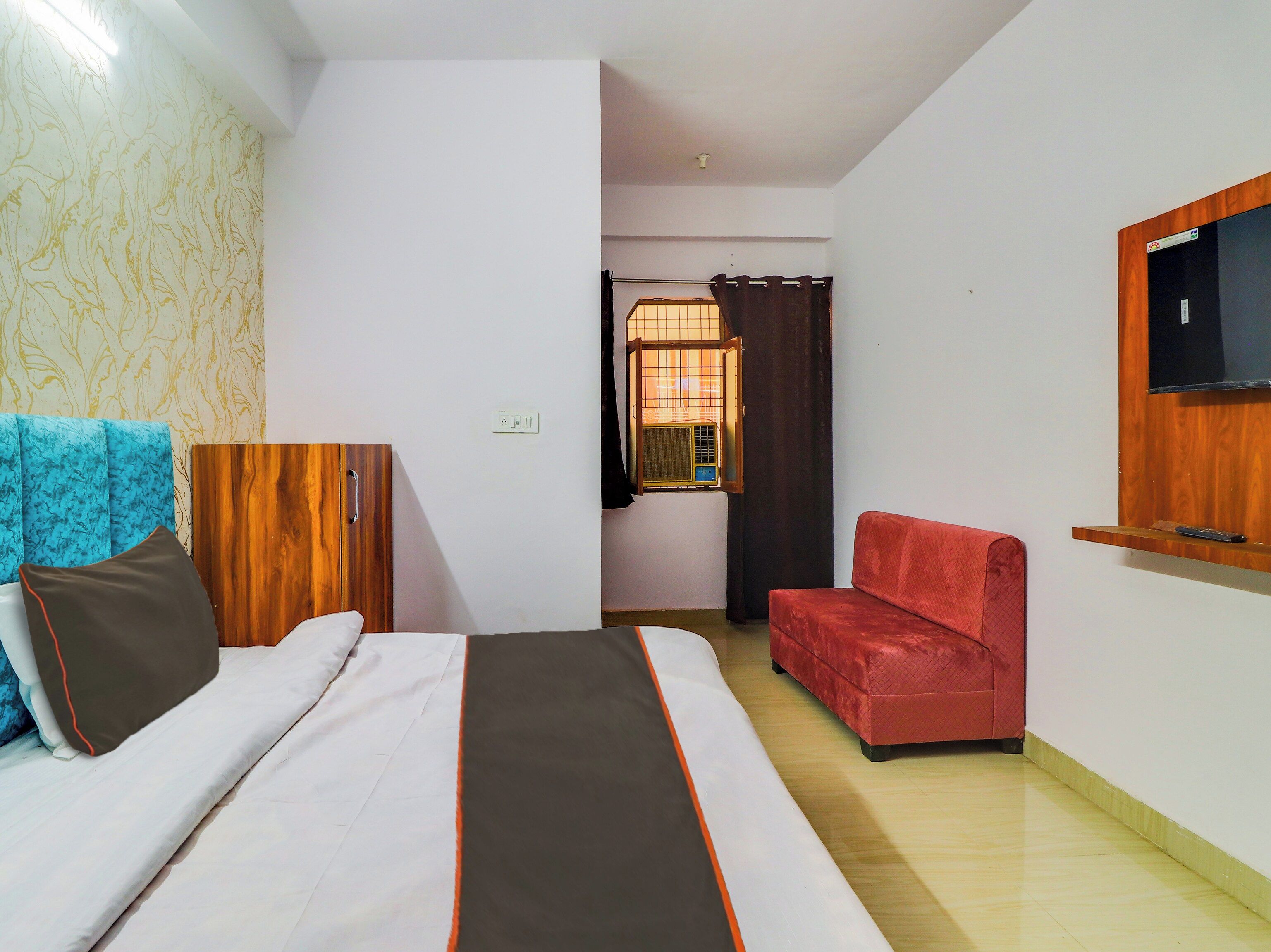 Une chambre d’hôtel avec un lit, un canapé rouge, un téléviseur fixé au mur et une fenêtre avec des rideaux.