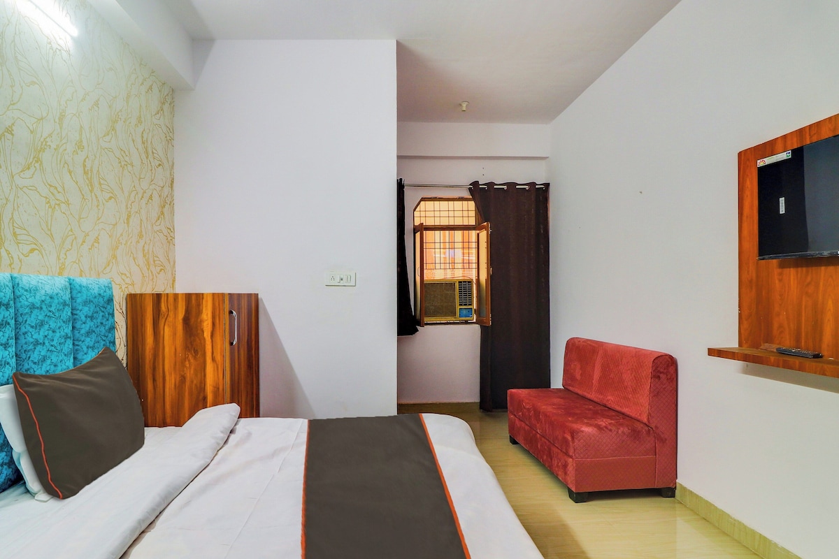 A hotel room with a bed, a red sofa, a TV mounted on the wall, and a window with curtains.