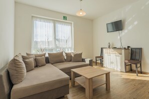 A modern living room with a sectional sofa, a wooden coffee table, a flat-screen TV mounted on the wall, and a small kitchenette area. - New Listing (Mainz)