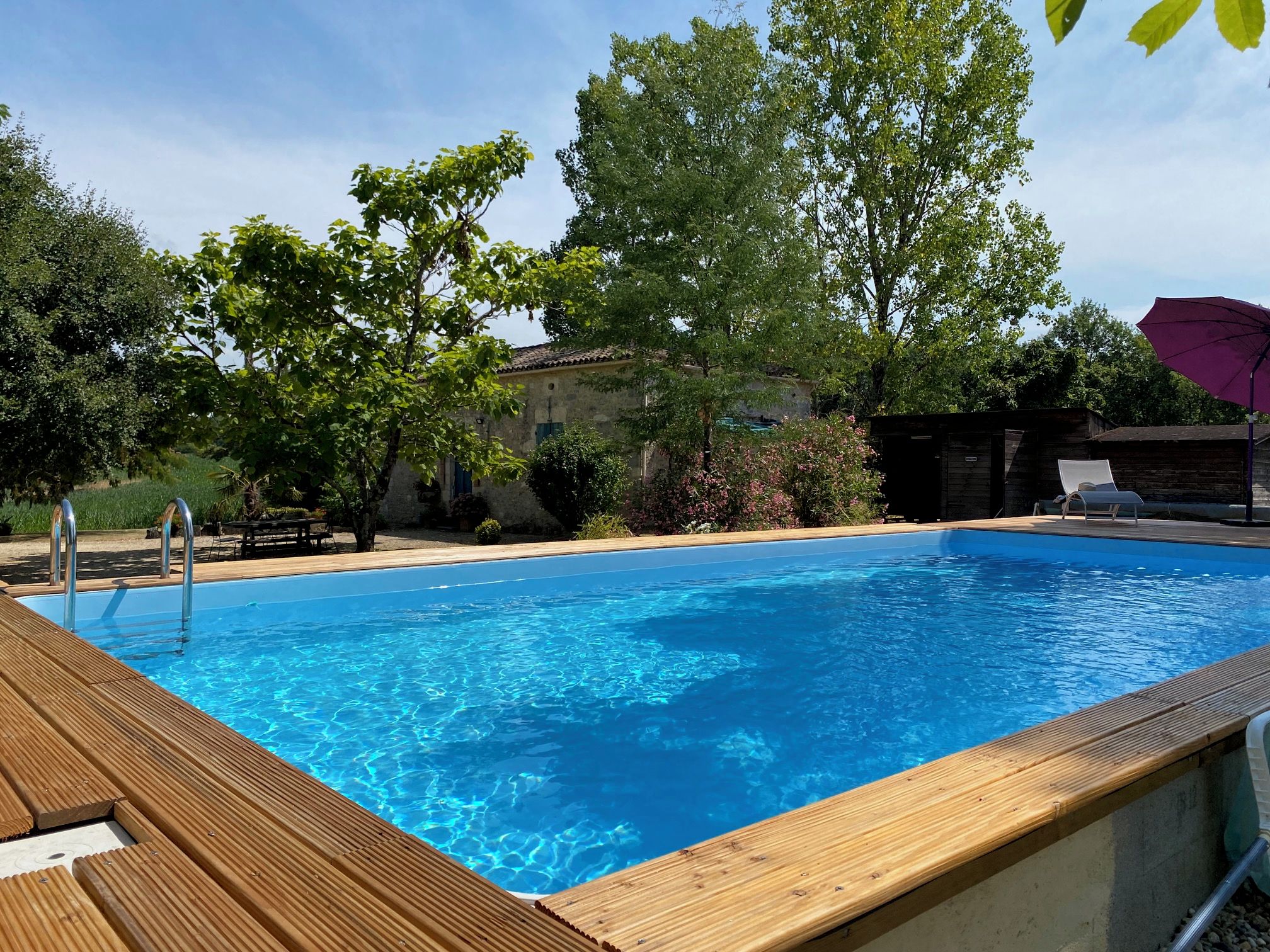 A rectangular swimming pool with a wooden deck edge, surrounded by trees and a stone building.