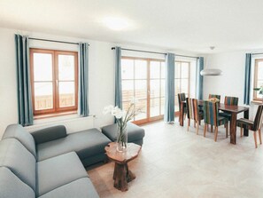 A modern living room with a sofa, dining table, chairs, and a view through large windows. - Luna Blanca - Apartment 2 (Schwangau)