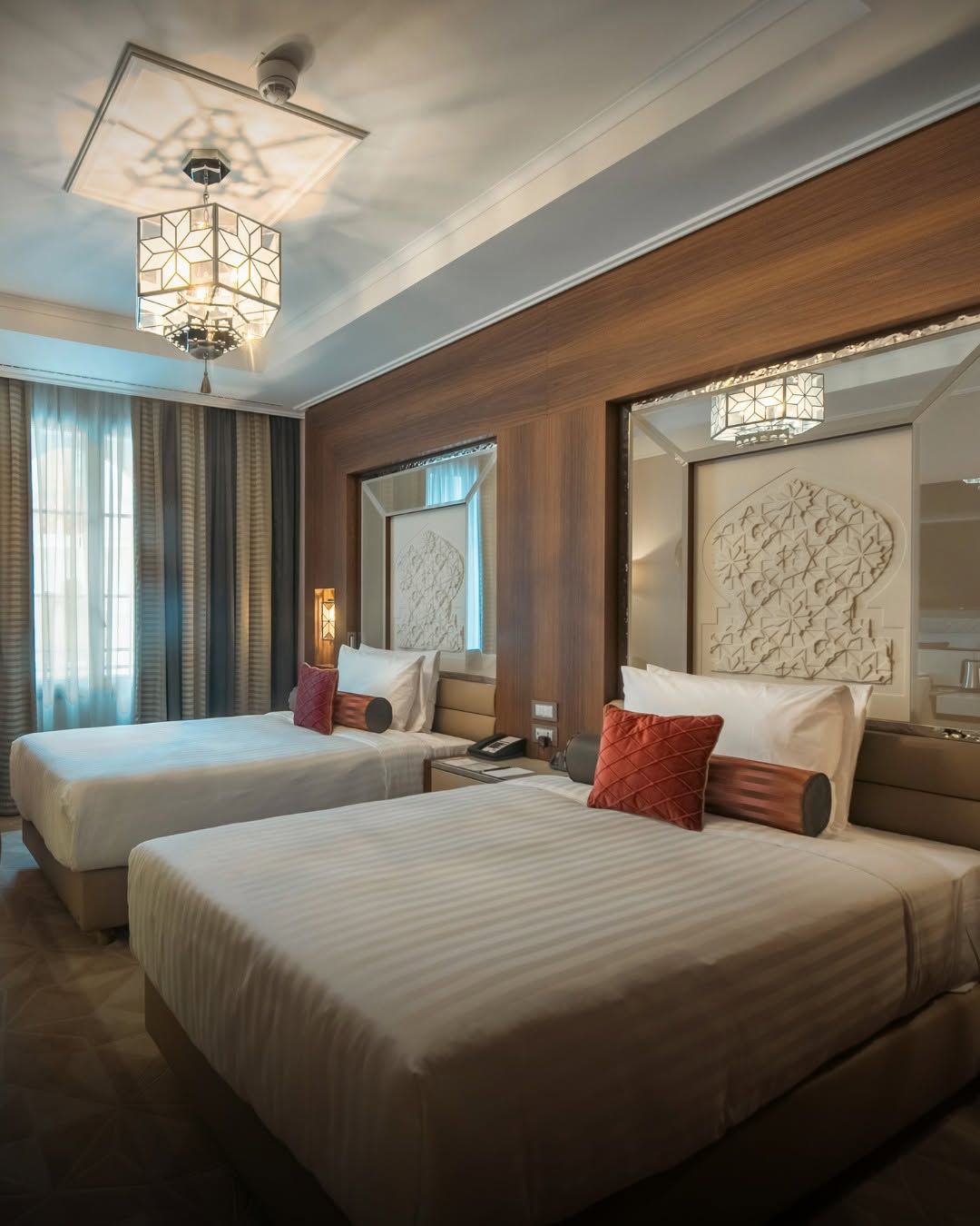 A hotel room with two beds, a large mirror, and a modern chandelier.