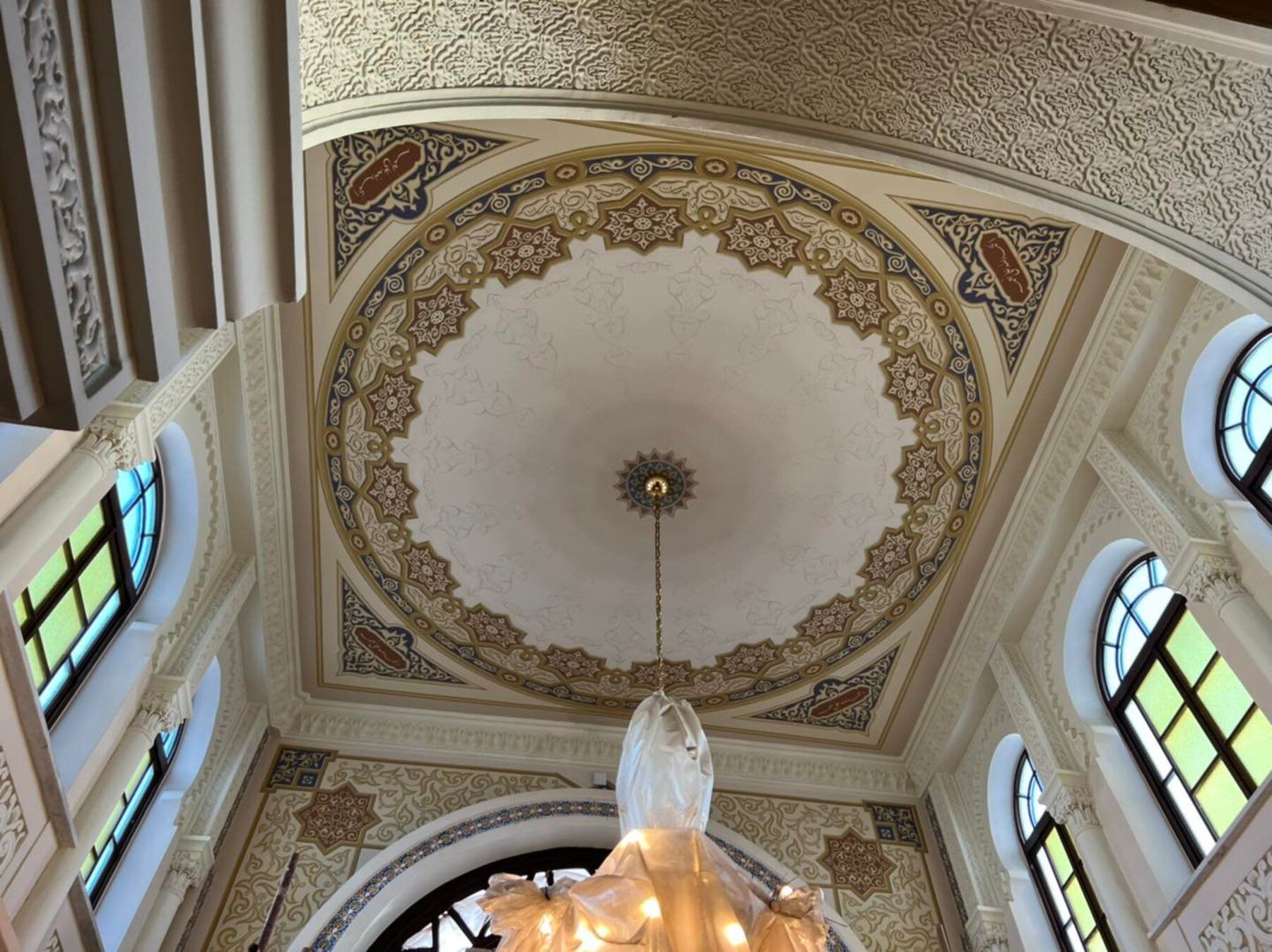 A high-ceilinged room with ornate ceiling patterns, a central chandelier, and multiple windows.