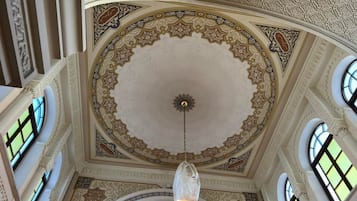A high-ceilinged room with ornate ceiling patterns, a central chandelier, and multiple windows.