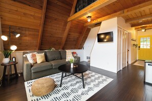 Un accogliente spazio abitativo mansardato con soffitto in legno, un divano, un tavolino da caffè e un poggiapiedi rotondo.