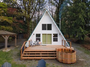 Un piccolo chalet a forma di A, bianco, con terrazza in legno, vasca idromassaggio e area salotto coperta.