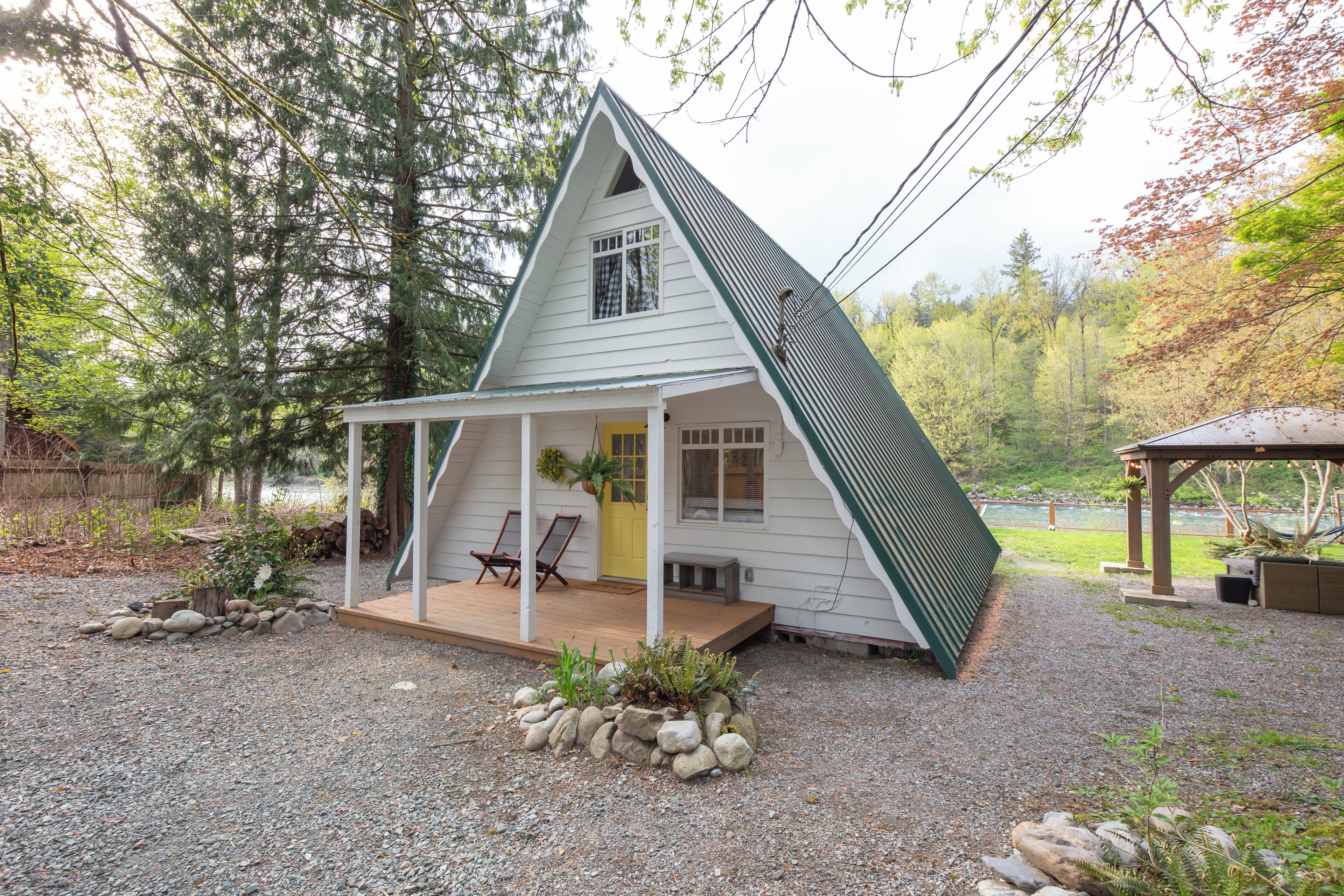 Ein kleines, weißes Einfamilienhaus mit Satteldach, einer Veranda mit zwei Stühlen und einem kleinen Garten mit Steinen und Pflanzen.
