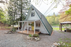 Une petite maison blanche, de plain-pied, avec un toit en pente, une véranda avec deux chaises et un petit jardin agrémenté de rochers et de plantes.