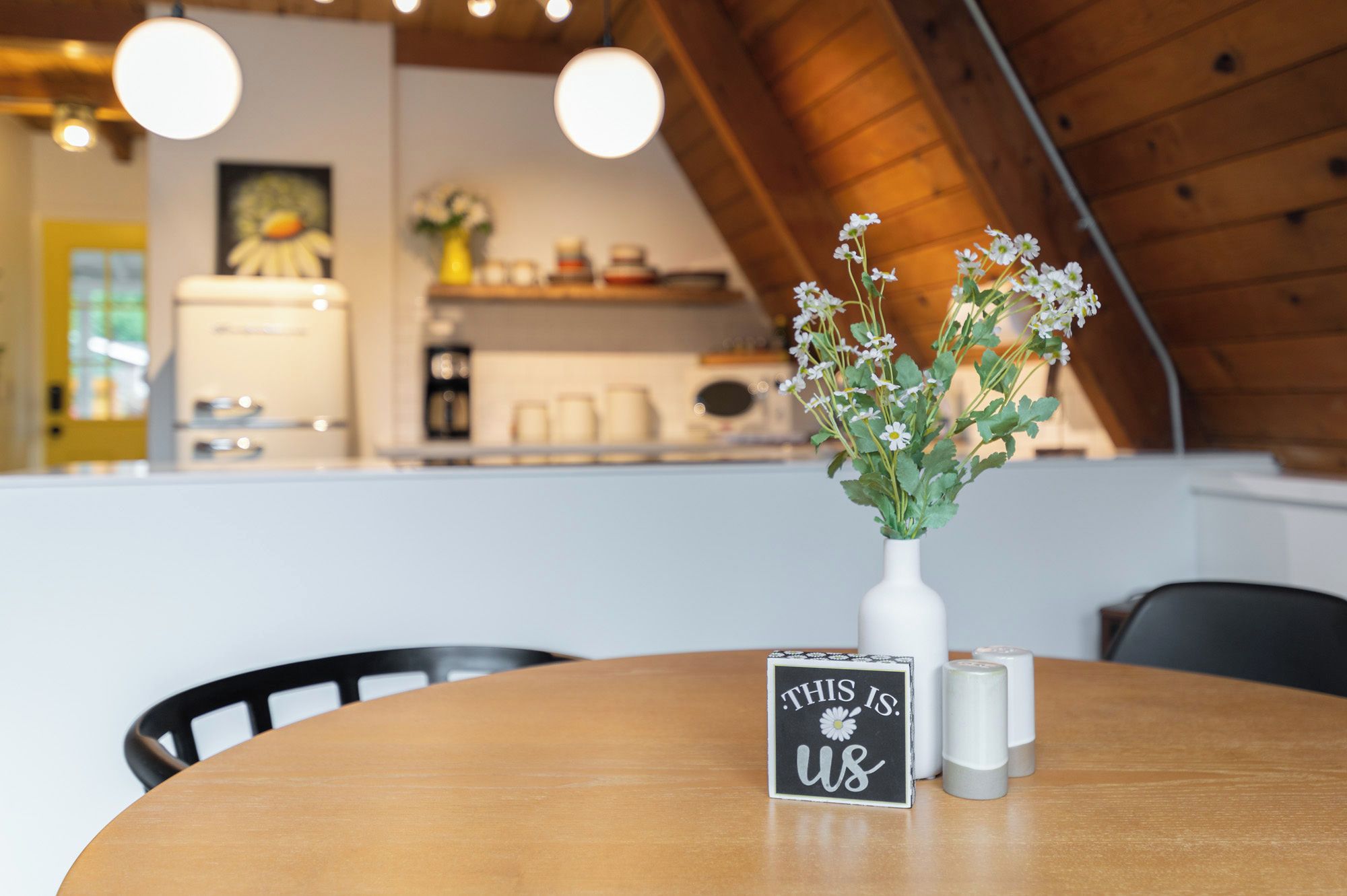 Una mesa de comedor con un jarrón blanco de flores, un cartel con texto y saleros.