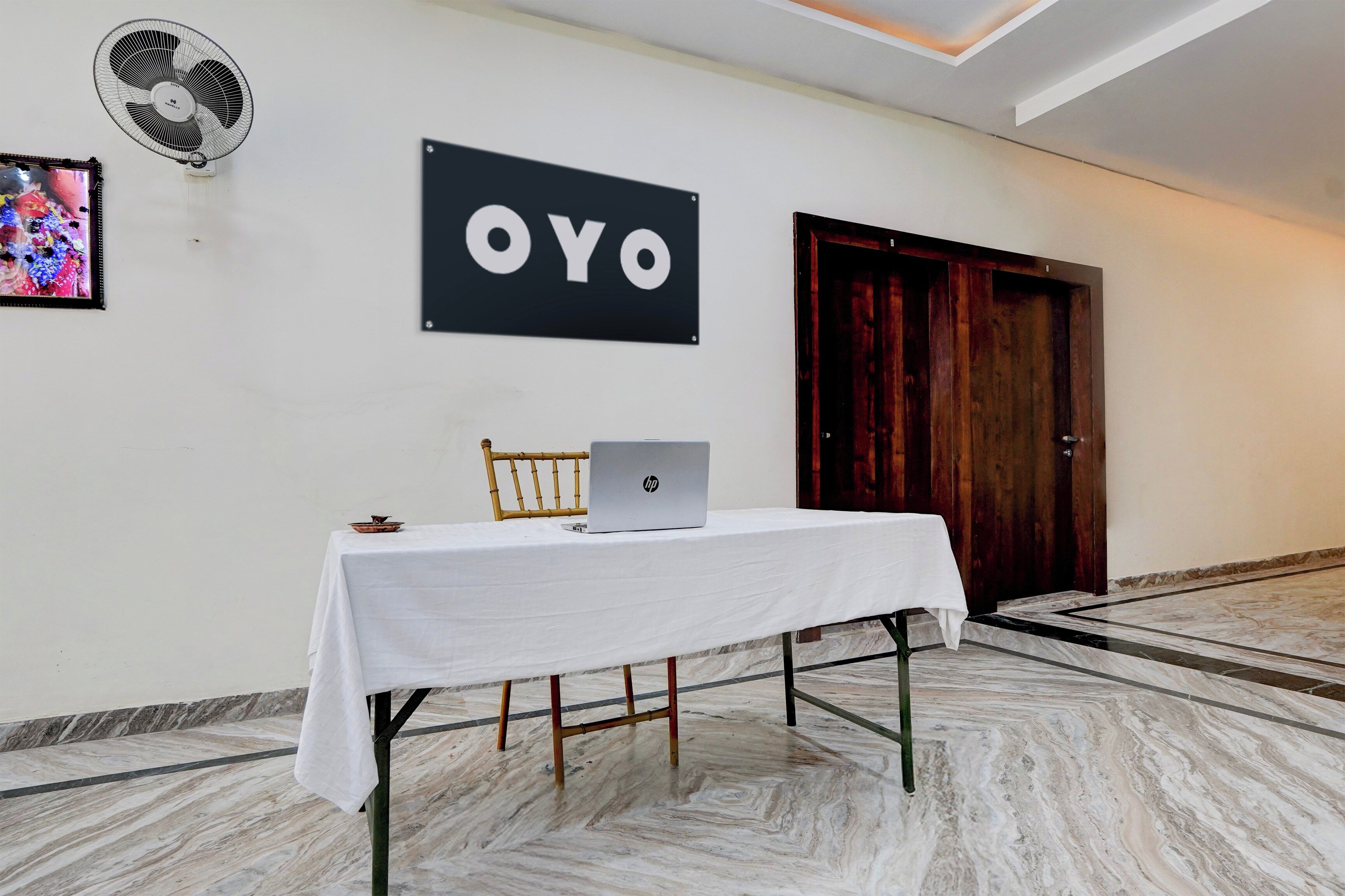 A hotel room with a white table, a laptop, a fan, and a door.