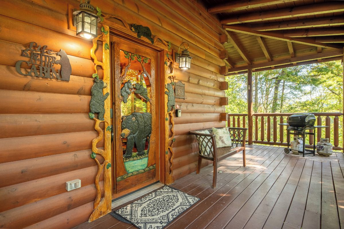 A wooden cabin with a decorative door, a porch with a grill, and a view of trees.