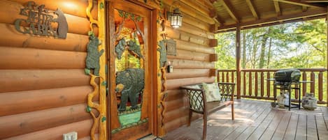 A wooden cabin with a decorative door, a porch with a grill, and a view of trees.