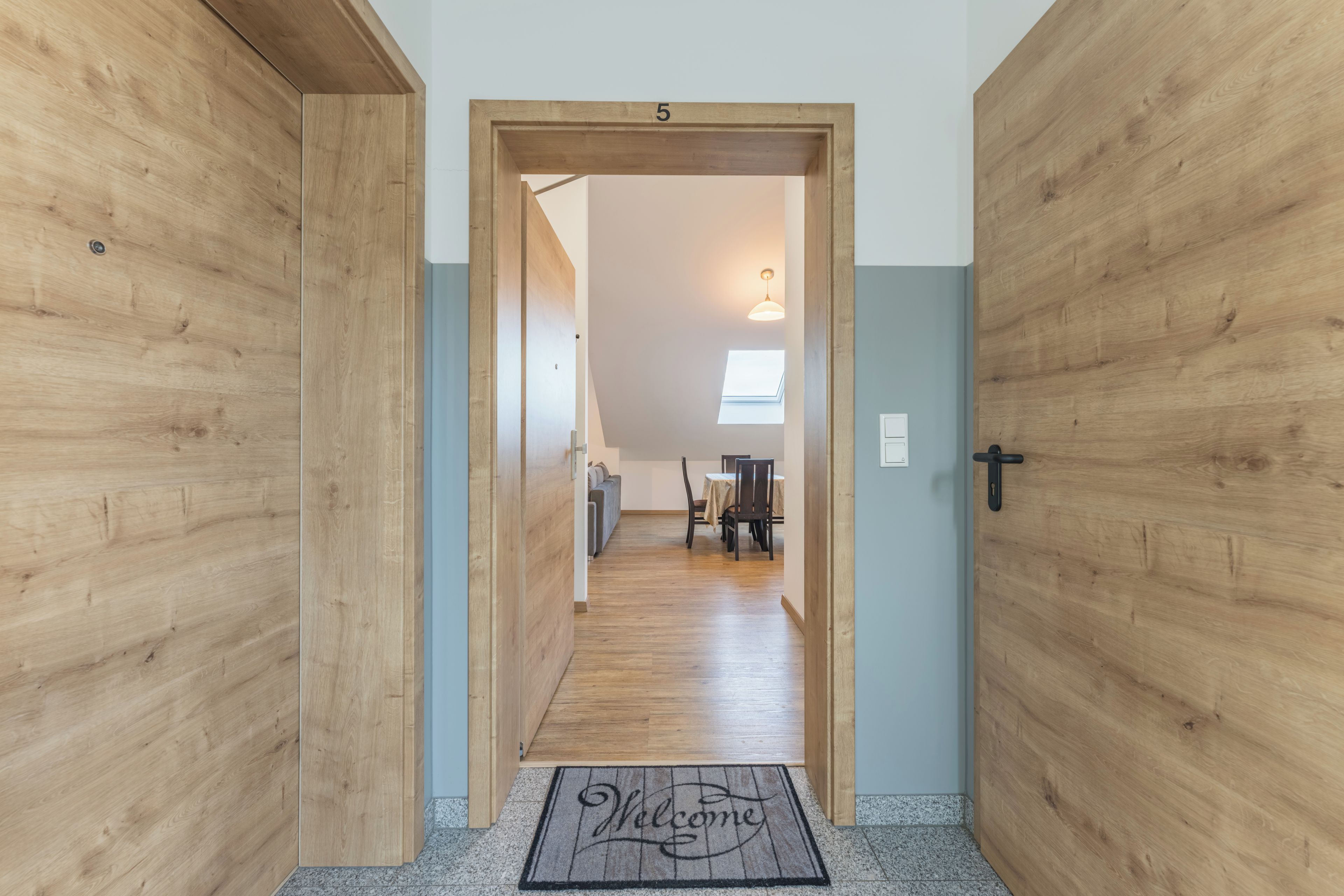 A hallway with wooden walls, a welcome mat, a door labeled '5', and a dining area in the background.