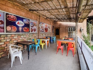 An outdoor seating area with colorful plastic chairs and tables, a brick wall, and posters advertising a hotel and restaurant. - Hotel O Anand Hotel Jodhpur (Jodhpur)