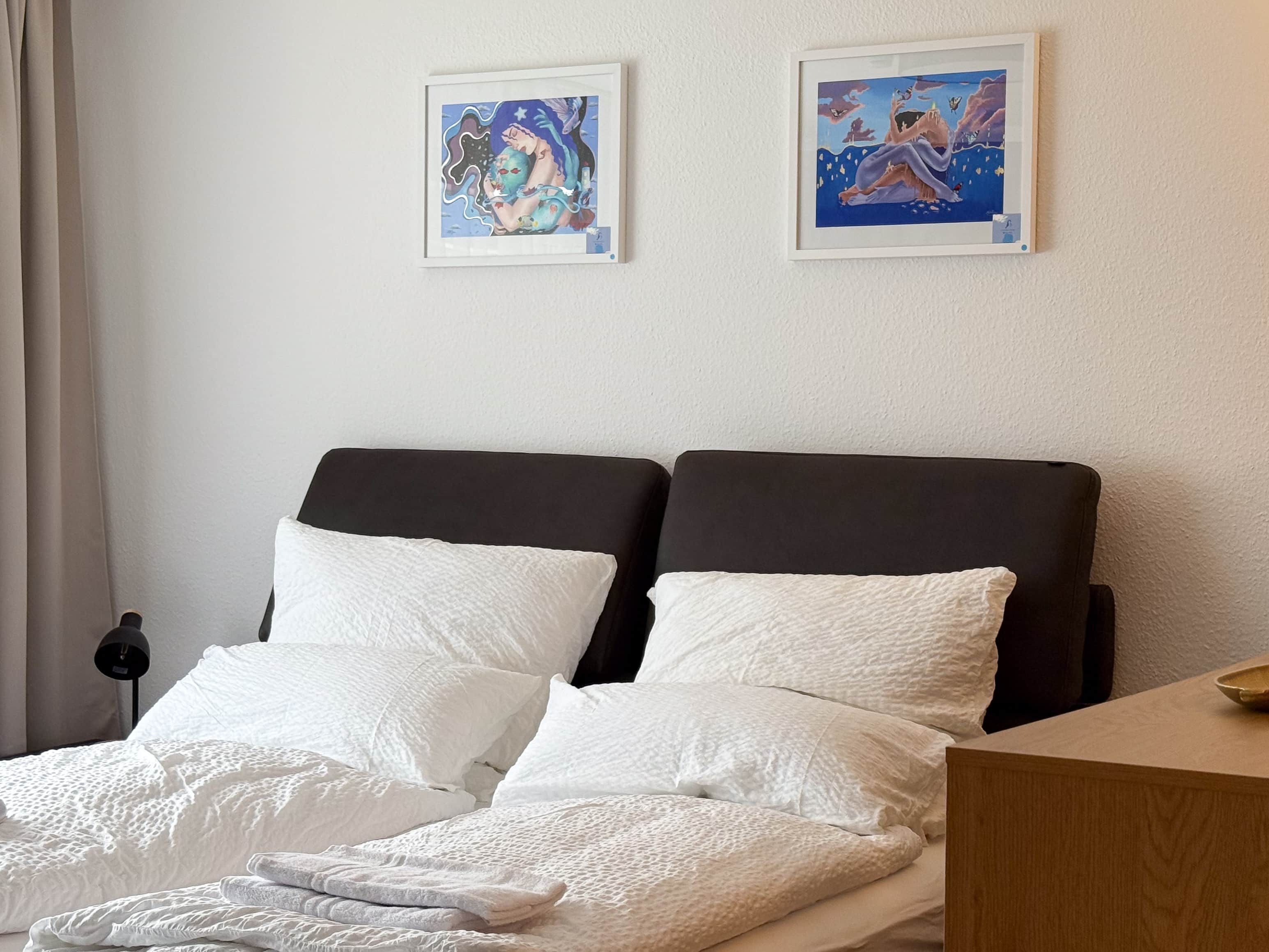 Ein Doppelbett mit weißer Bettwäsche, zwei gerahmte Kunstwerke an der Wand und ein Nachttisch mit einer Lampe.