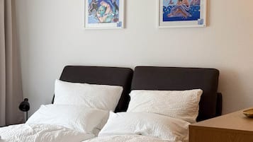 Ein Doppelbett mit weißer Bettwäsche, zwei gerahmte Kunstwerke an der Wand und ein Nachttisch mit einer Lampe.