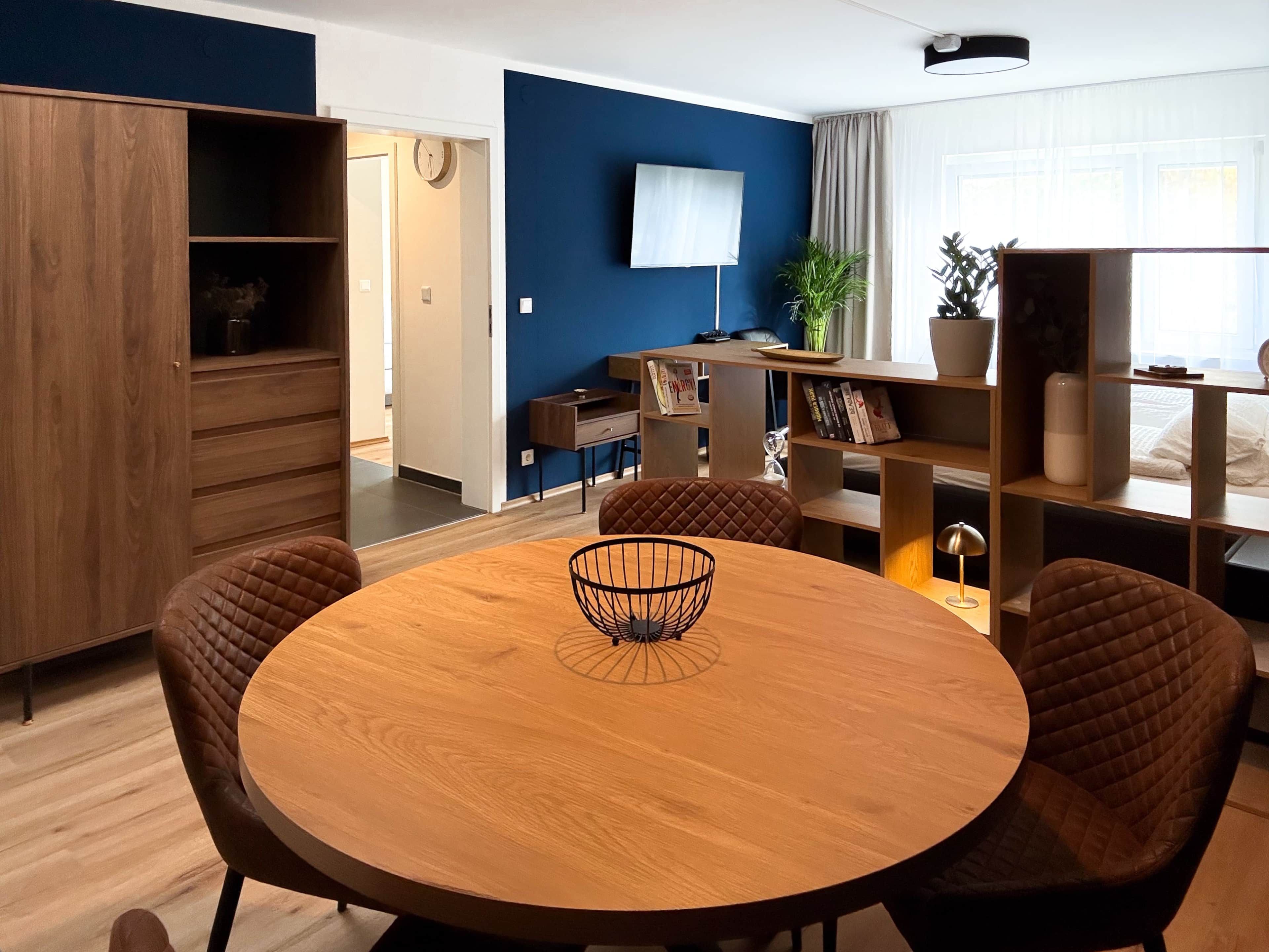 A modern living room with a round wooden table, two brown chairs, a TV mounted on the wall, and a bookshelf.