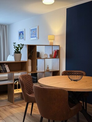A modern living room with a round wooden table, two cushioned chairs, a bookshelf, a potted plant, and a wall-mounted lamp.