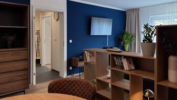 A modern living room with a wooden table, a brown cushioned chair, a bookshelf, a flat-screen TV, and a potted plant.
