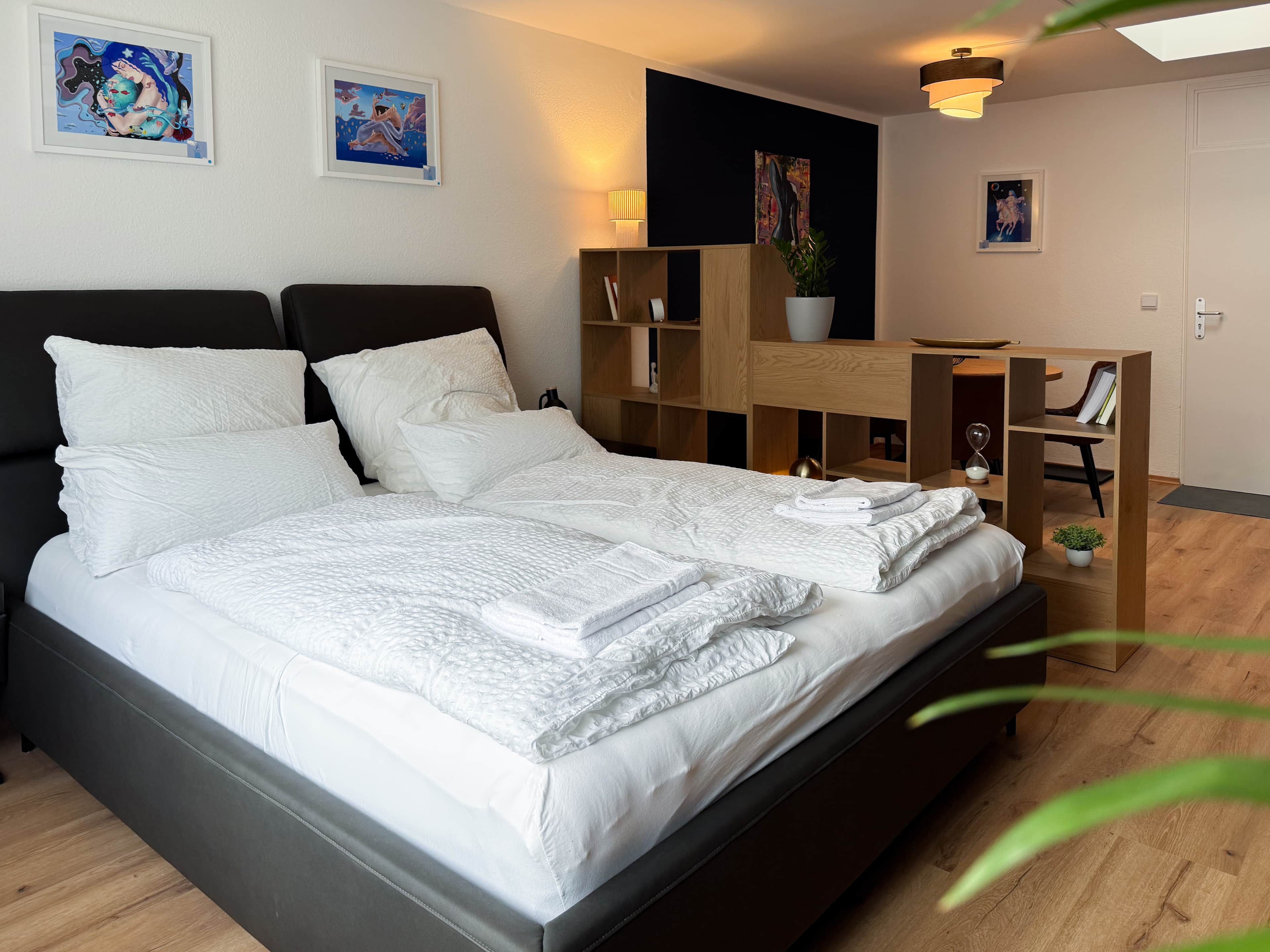 A neatly made bed with white linens and pillows, a wooden nightstand with a lamp, and a wall with two framed artworks.
