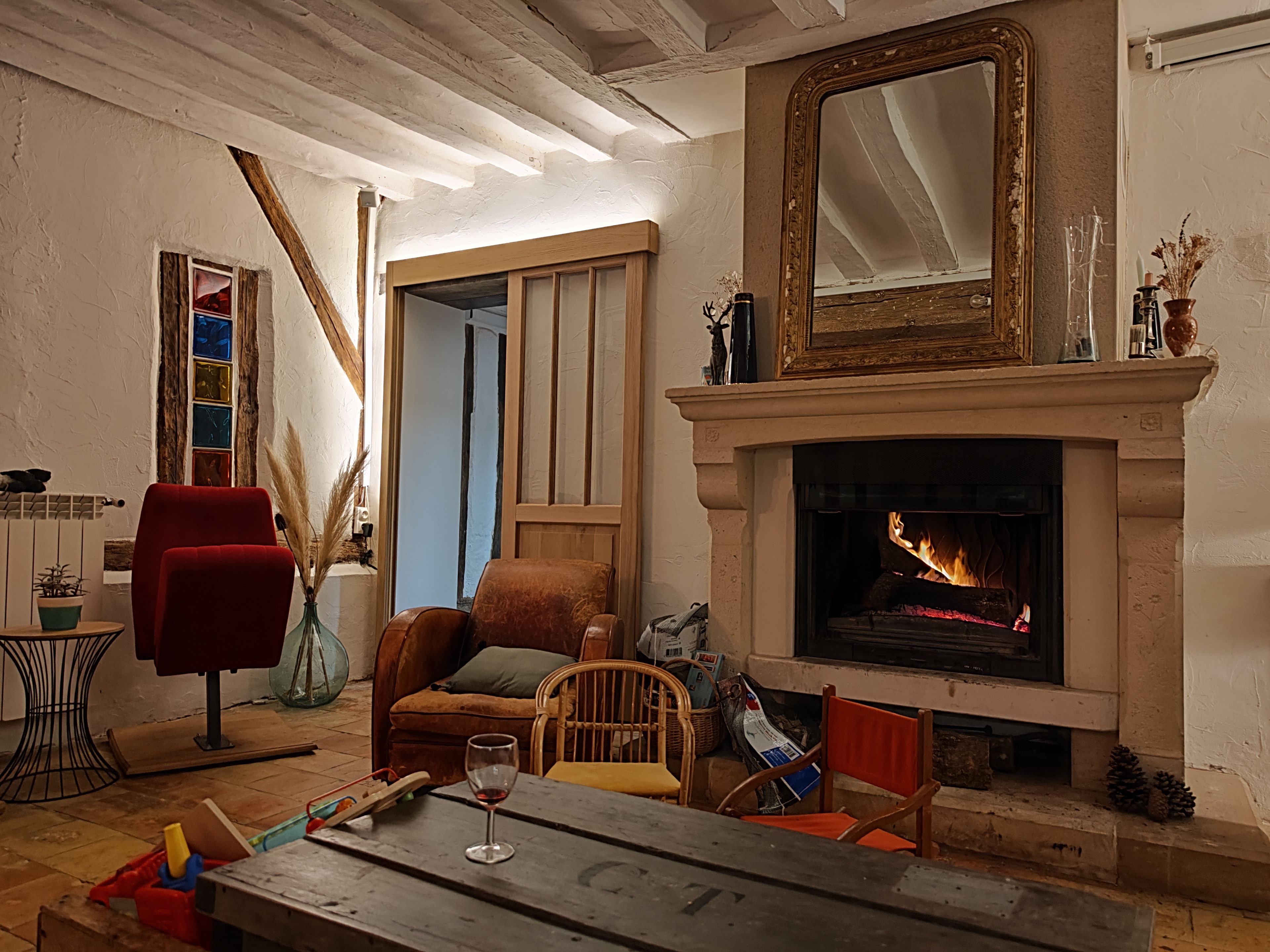 A cozy living room with a fireplace, a red armchair, a wooden table, and a large mirror above the mantel.